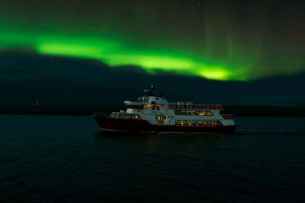 Elding's boat lit up on the dark water of Faxaflói Bay with a band of green aurora above it