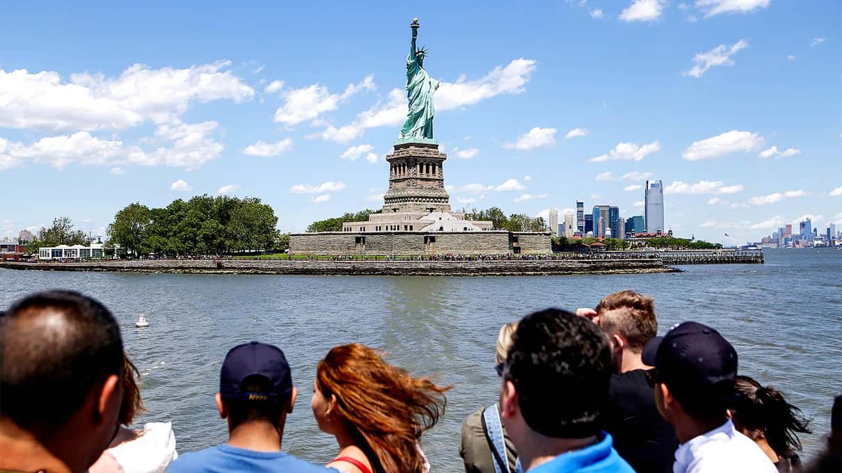 Ferry passengers crowding the rail as the boat approaches Liberty Island under a blue sky