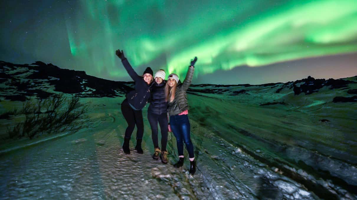 Three people raising their arms on snowy ground under a wide green aurora on an Arctic Adventures night