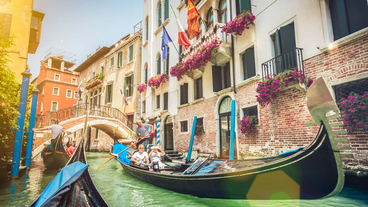Gondole dans un canal calme bordé de façades fleuries et de drapeaux, avec un petit pont en arrière-plan