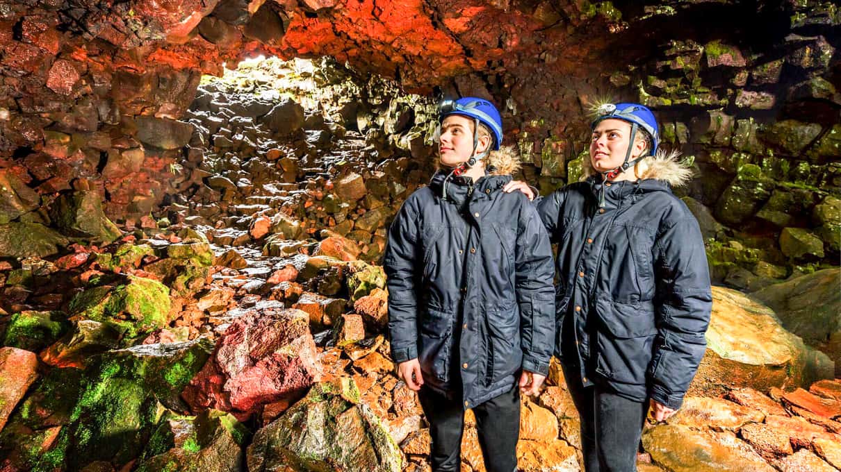 Two visitors in helmets inside the Raufarhólshellir lava cave with mineral-colored walls