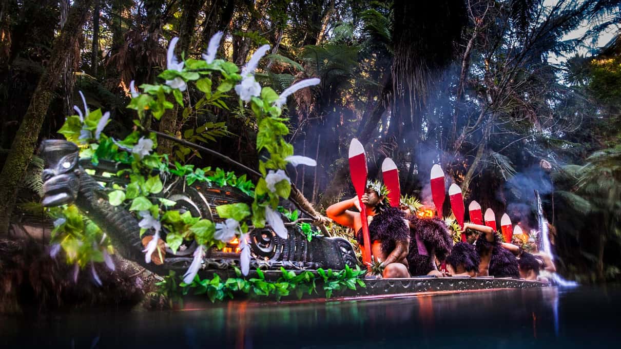 Warriors with red and white paddles in a carved waka decked with ferns and feathers, lit by torches at night