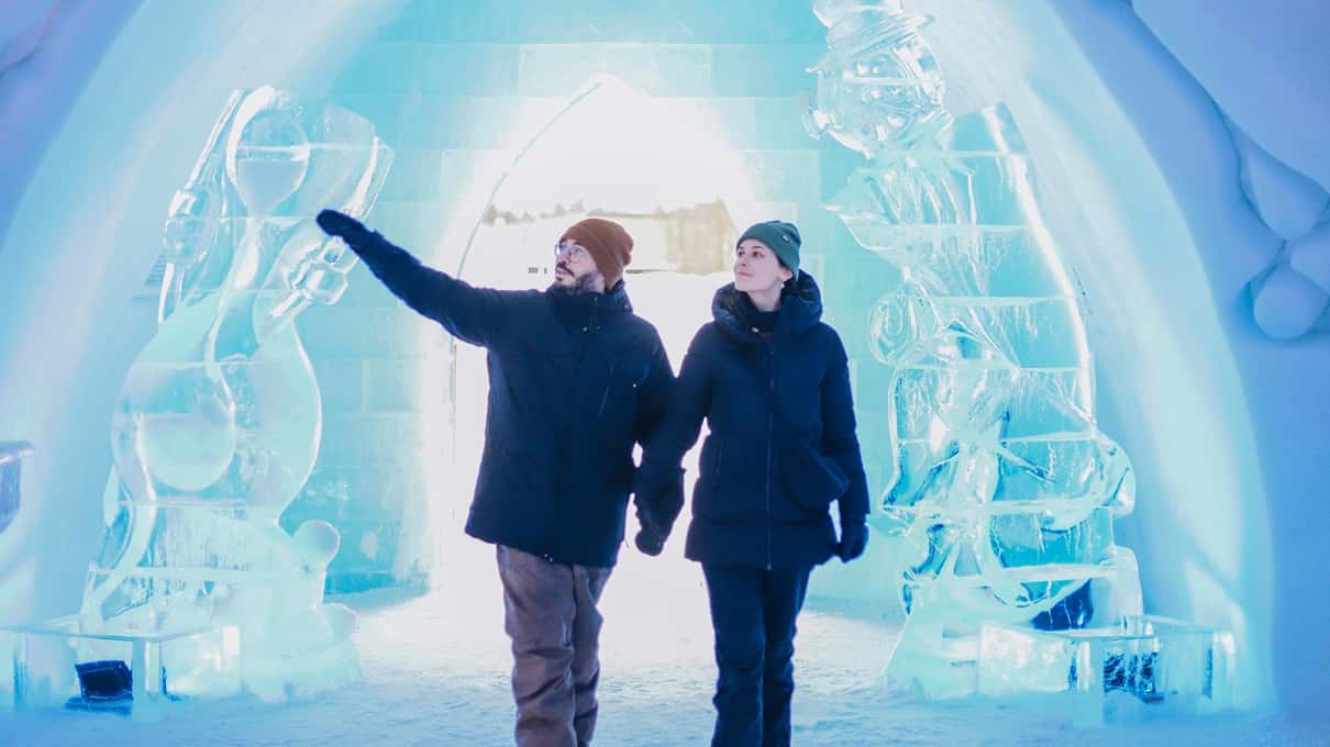 Two visitors in parkas pointing at blue-lit ice sculptures under a snow arch inside the Hôtel de Glace