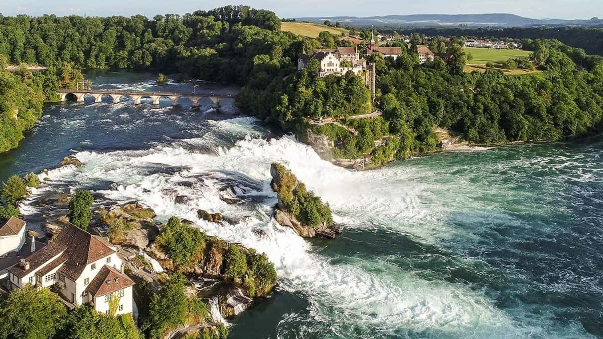 The Rhine Falls pouring white over rocks below Laufen Castle on its wooded cliff