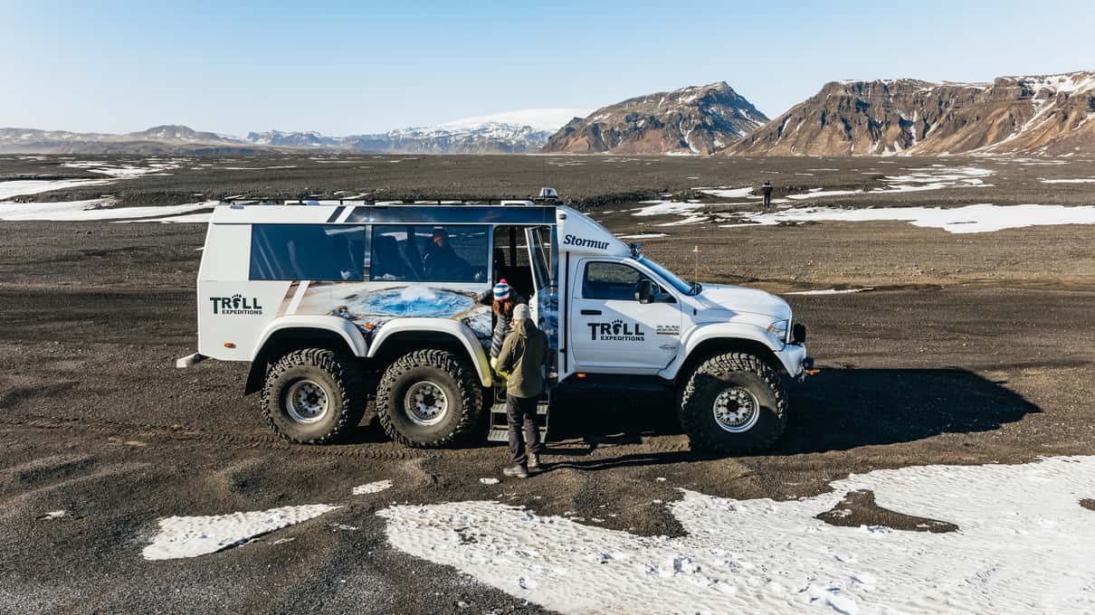 Un Super Jeep blanc de Troll.is dans lequel montent des passagers, sur du sable noir parsemé de neige, avec des montagnes tabulaires en arrière-plan