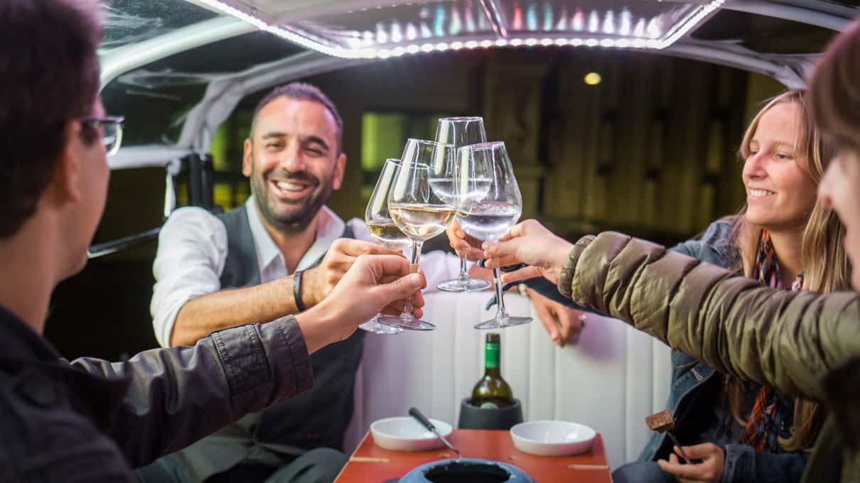 Two guests clinking glasses of white wine inside a covered e-tuk-tuk at night with a fondue pot on the table
