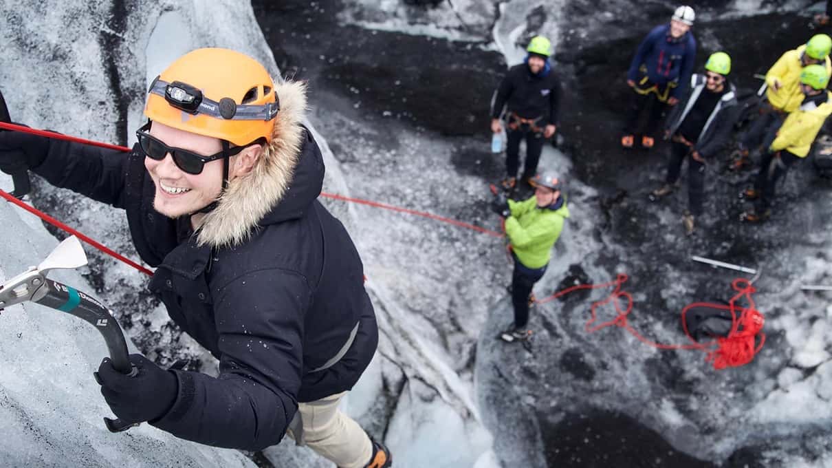 A climber in an orange helmet halfway up a glacier ice wall while a guide holds the rope below