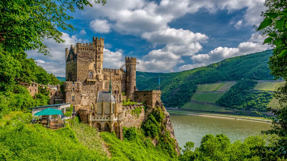 A medieval castle with square towers on a wooded rock above the Rhine, vineyards on the slopes across the water