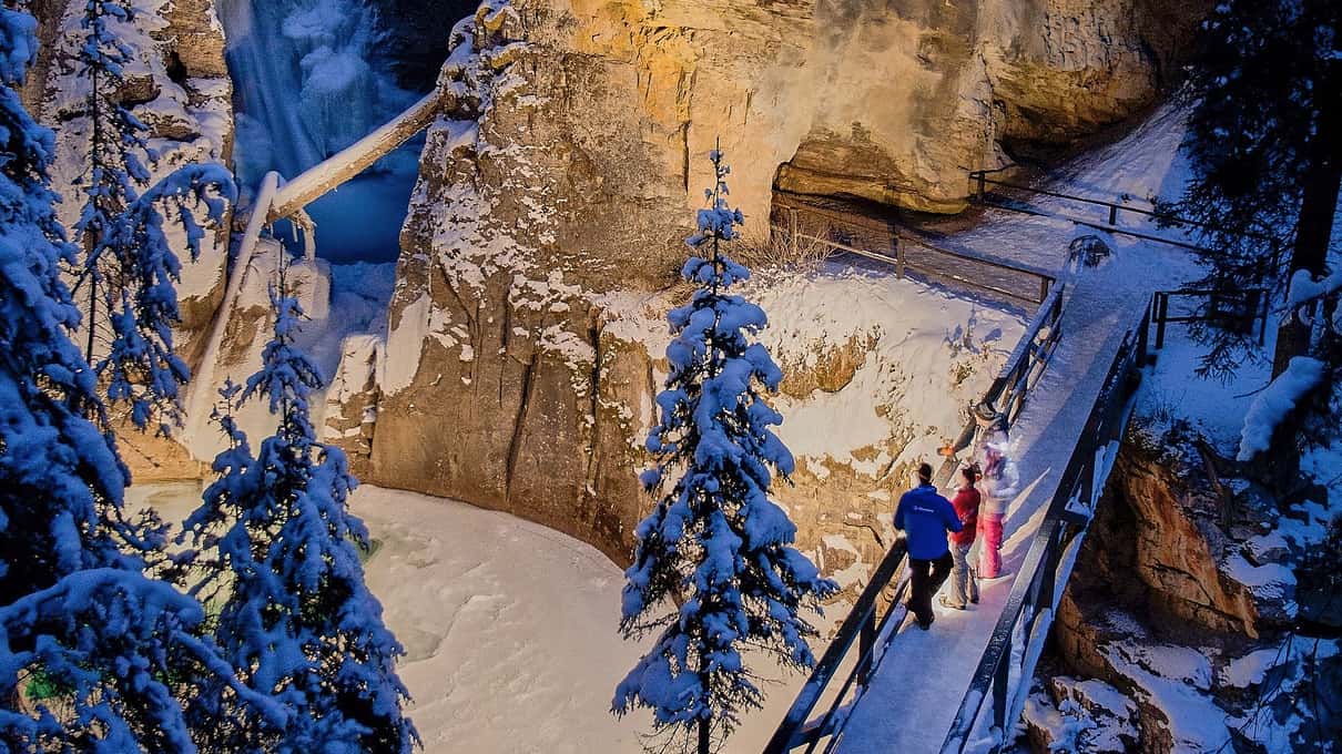 Two hikers on a lit steel catwalk between snowy canyon walls at dusk in Johnston Canyon