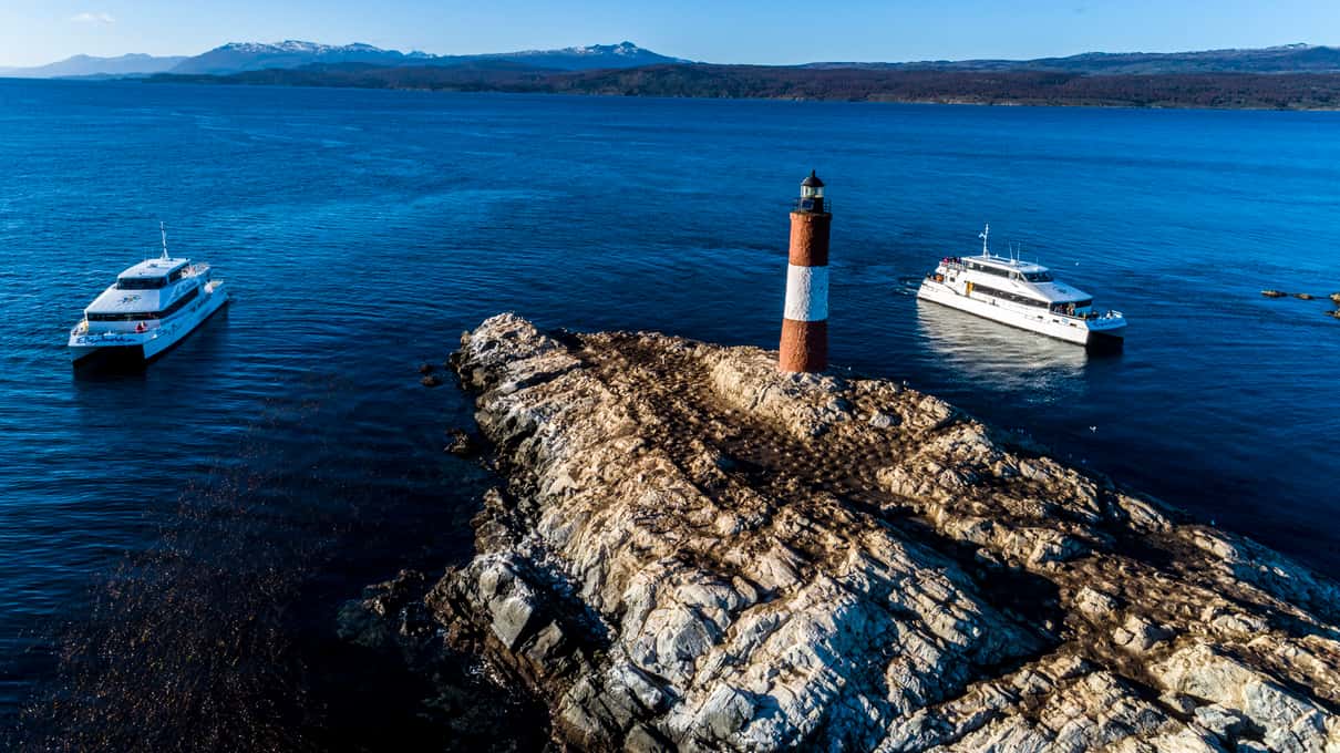 Aerial view of the red-and-white Les Eclaireurs lighthouse on its rock with boats on the deep blue Beagle Channel