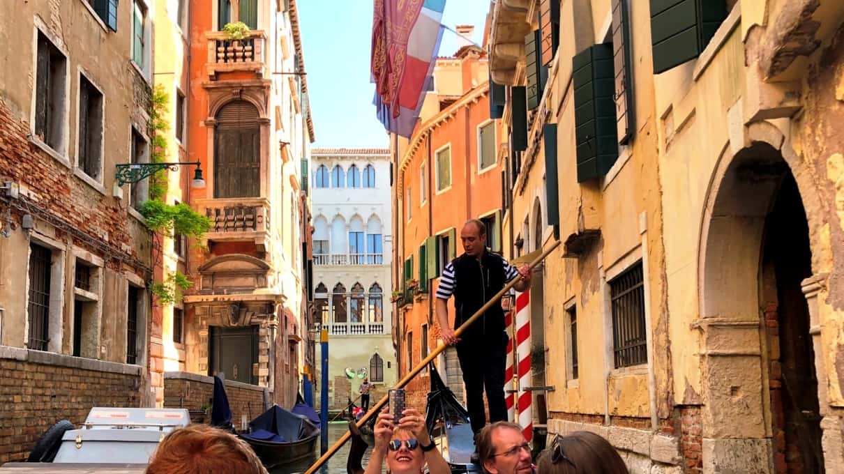 Gondolier w kamizelce wiosłuje wąskim kanałem między kolorowymi kamienicami z flagami, pasażerowie robią zdjęcia