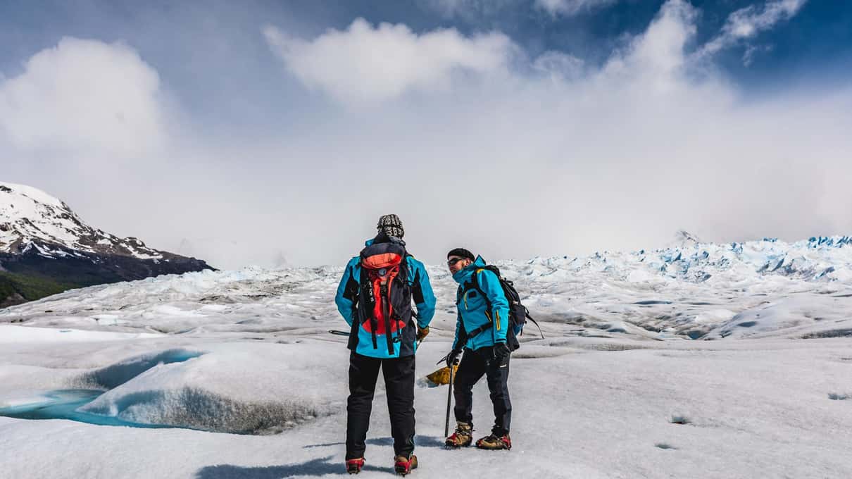 Two hikers wearing crampons standing on Perito Moreno ice beside a small blue meltwater pool