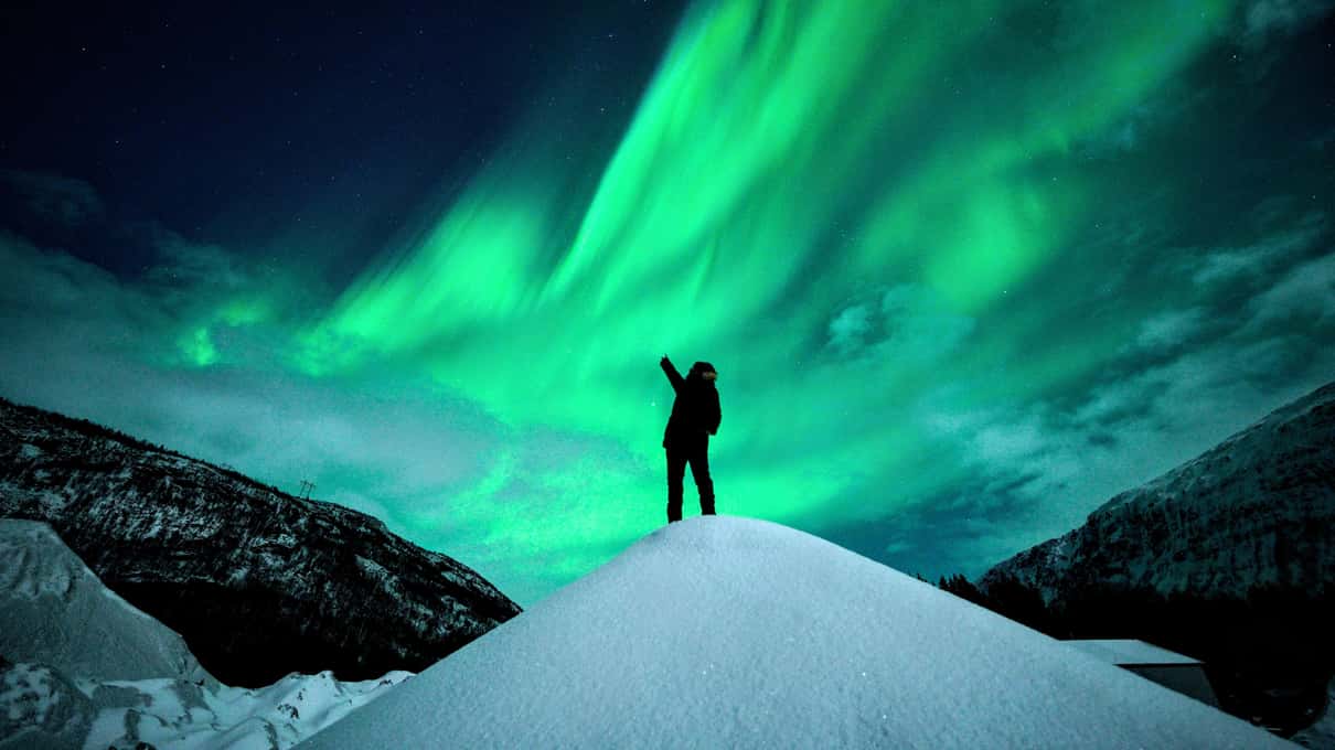 A lone figure with one arm raised on a snowy hilltop beneath a bright green aurora