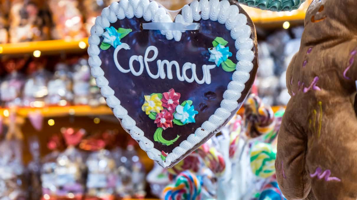 A large iced gingerbread heart reading Colmar hanging at a Christmas market stall full of sweets