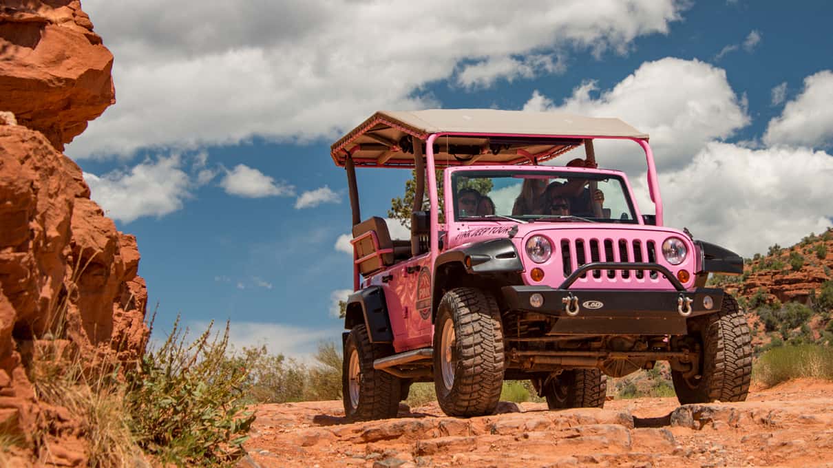 A pink open-top Jeep Wrangler parked on red slickrock high above the Sedona desert with buttes behind