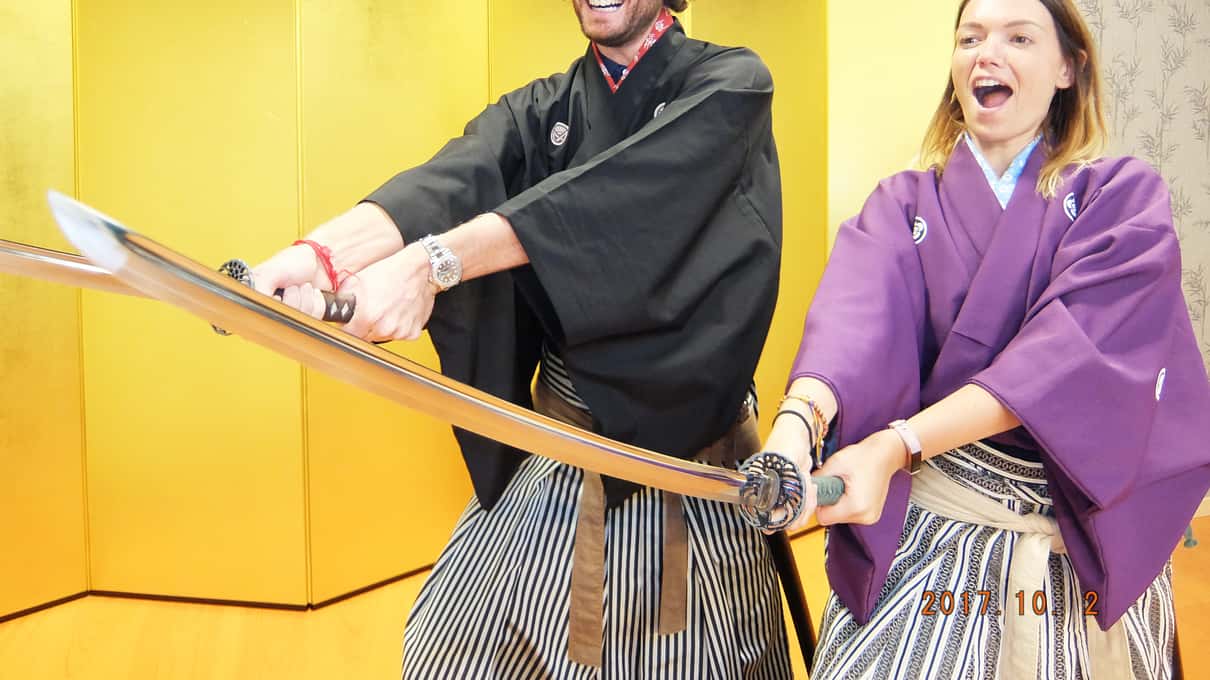 Smějící se muž a žena v hakama pózují s meči před zlatou skládací zástěnou v Kembu Theater