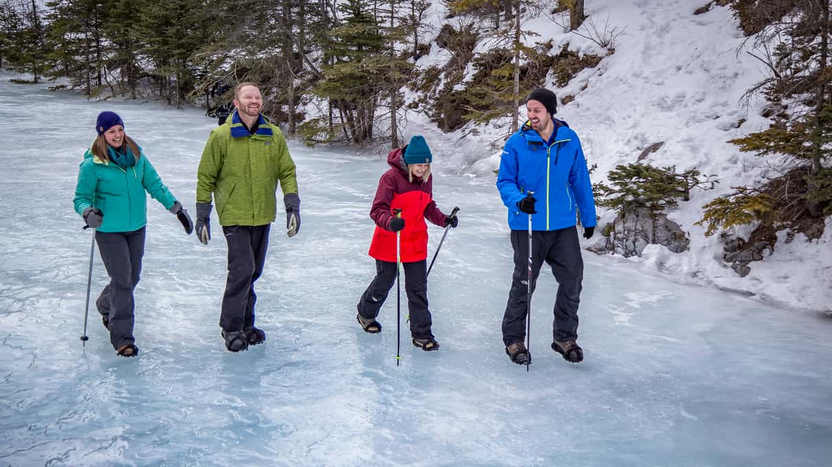 Four walkers with hiking poles in winter jackets and hats crossing a wide sheet of pale blue ice