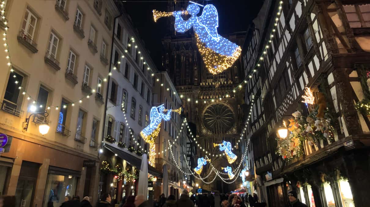 A narrow lane at night strung with gold lights and glowing angel figures, leading toward the lit cathedral facade