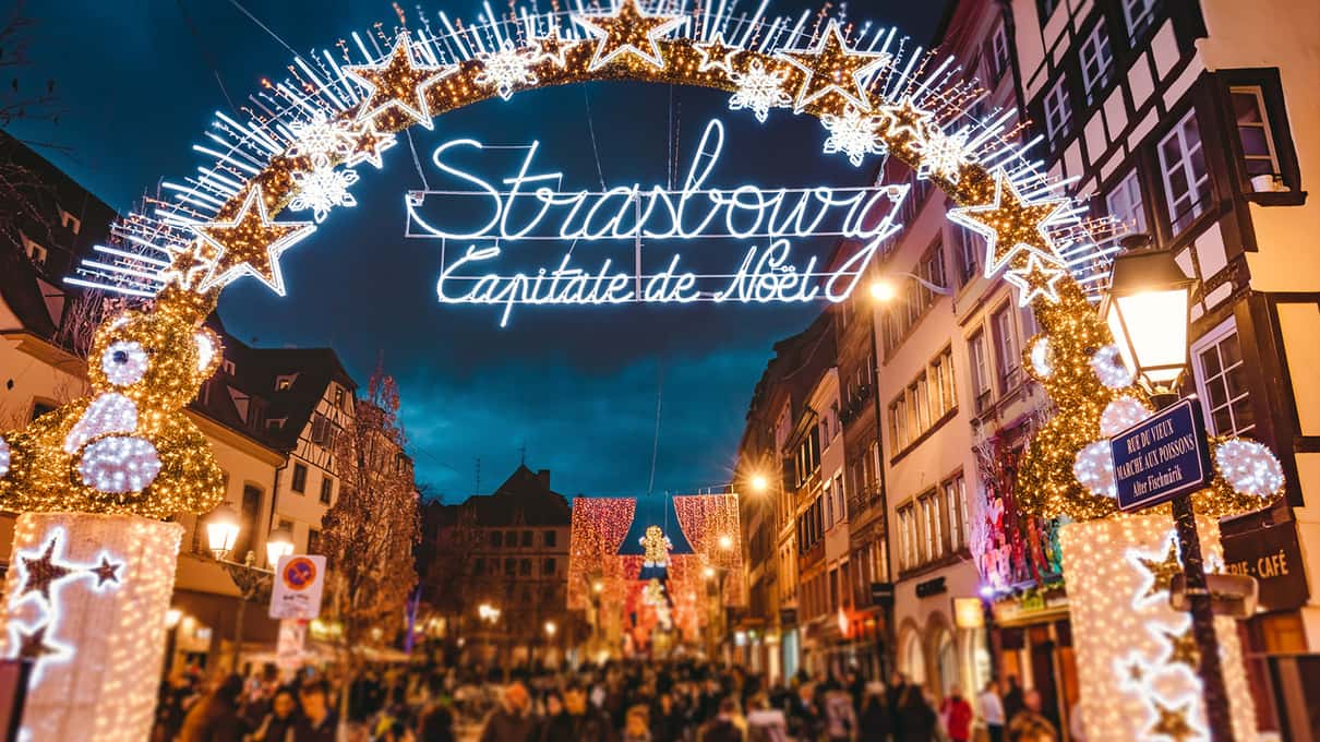 A star-shaped arch of lights reading Strasbourg Capitale de Noël over a busy street at night, flanked by two glowing gold teddy bears