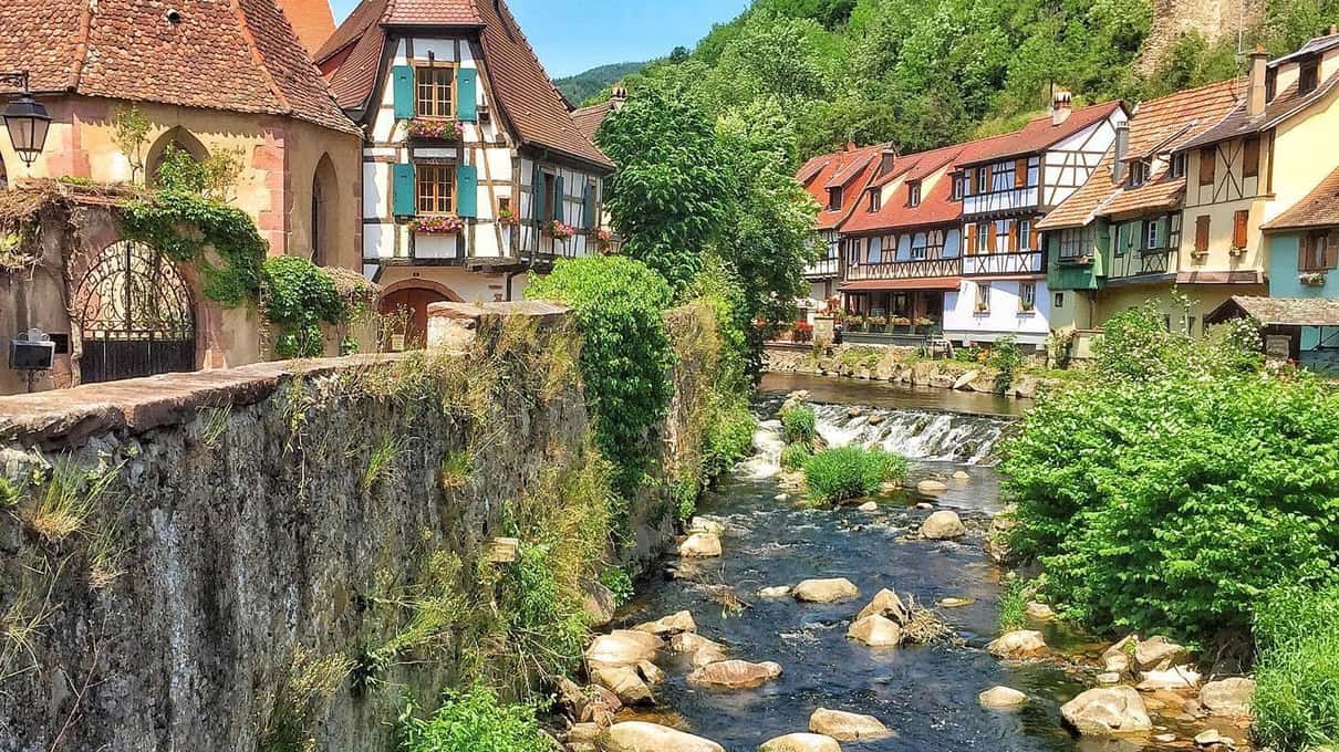Colourful half-timbered houses above a rocky stream in Kaysersberg, with a stone bridge and green hills behind