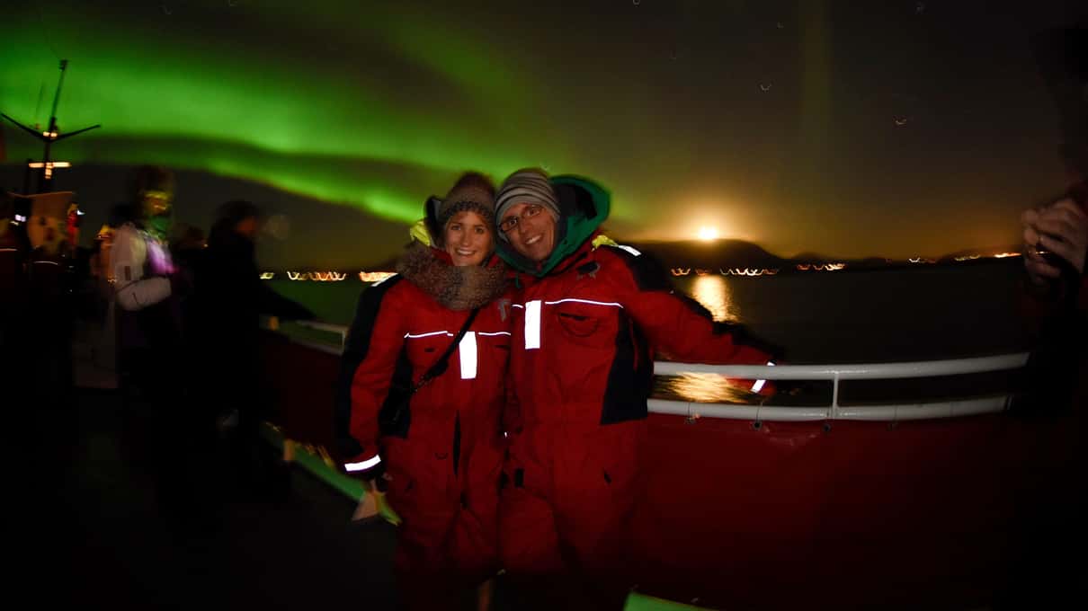 Guests wearing red Elding overalls standing on the dark deck of a boat under a green aurora