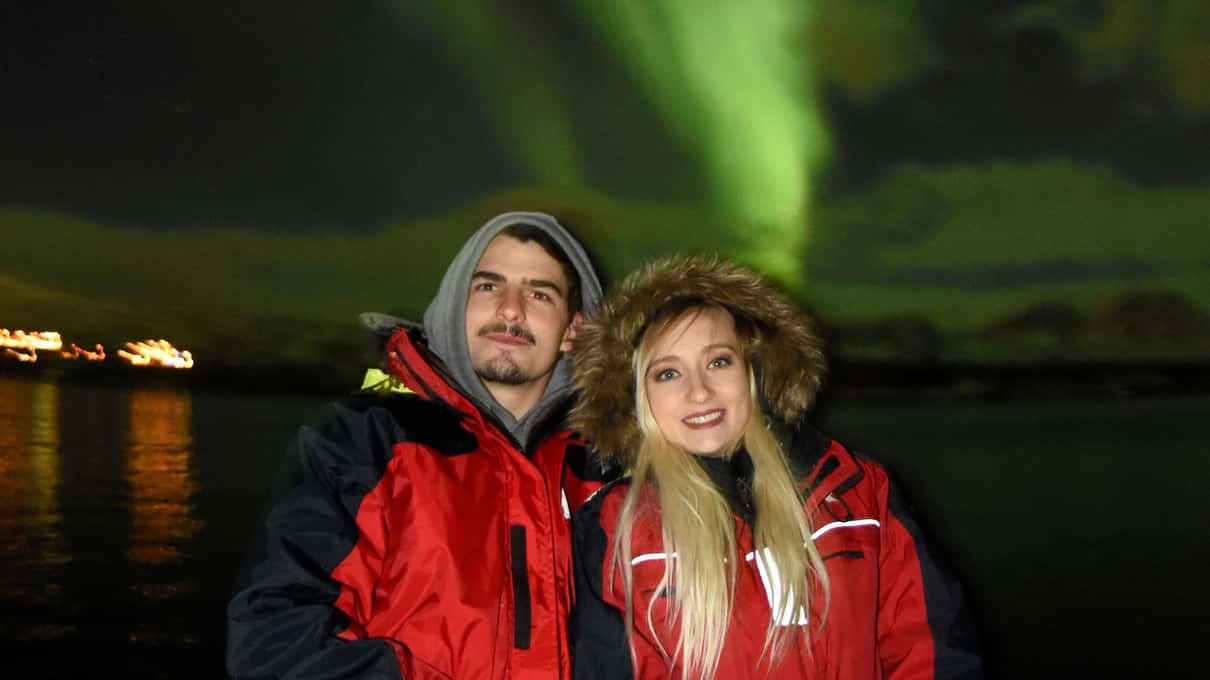 A smiling couple in red Elding overalls on the deck at night with a green aurora over the bay behind them