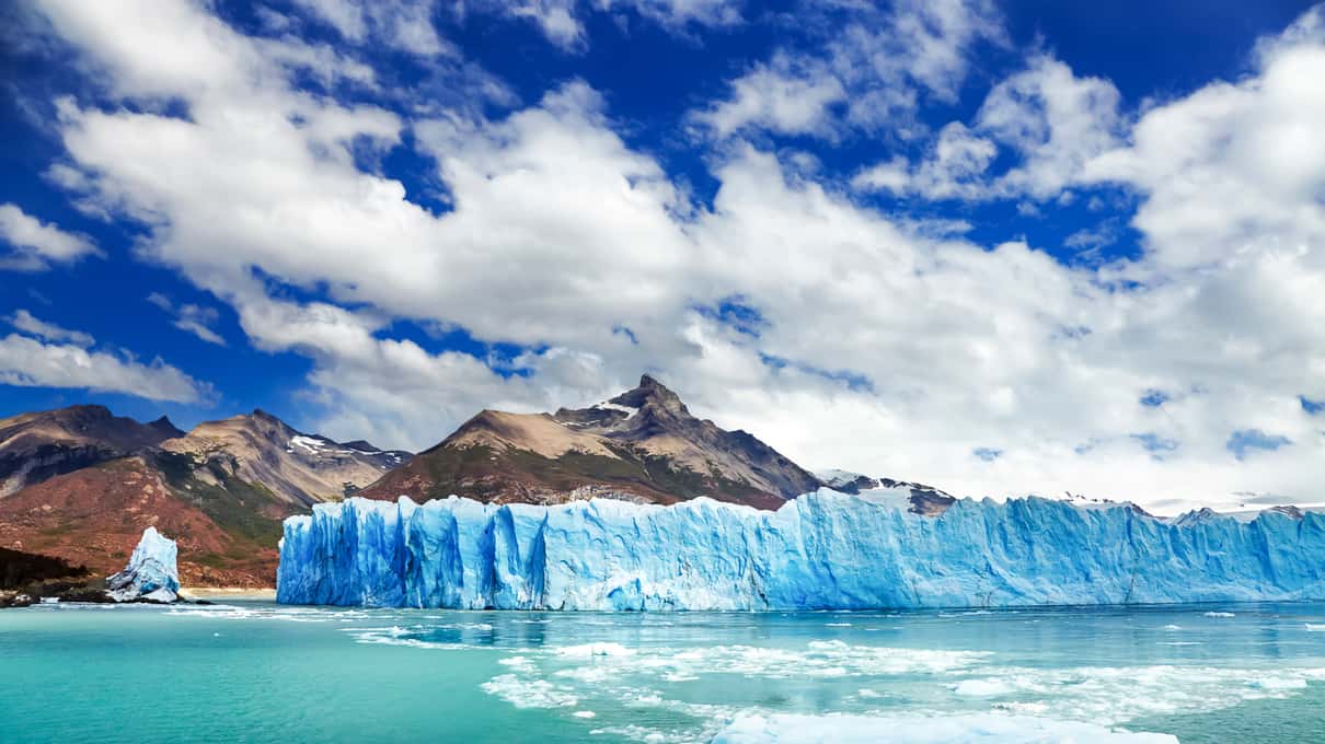 The front of Perito Moreno Glacier rising over turquoise lake water, with mountains under a blue sky
