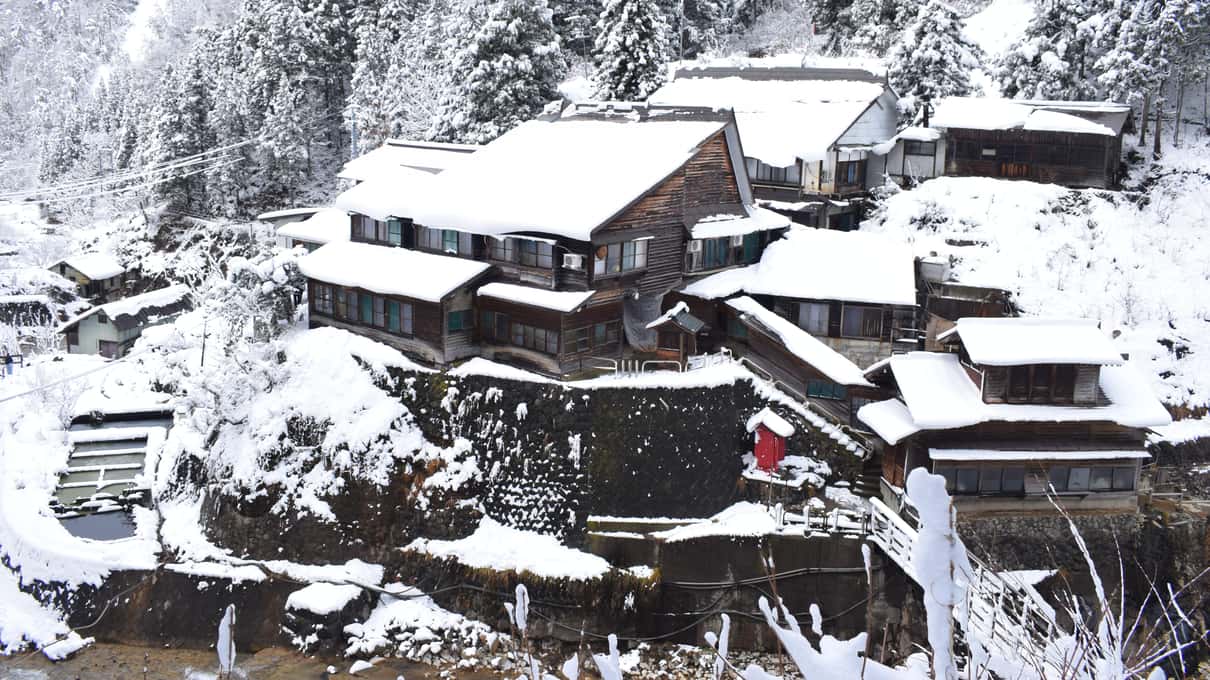 Snow-covered wooden buildings at the bottom of the steep, forested Jigokudani valley in winter