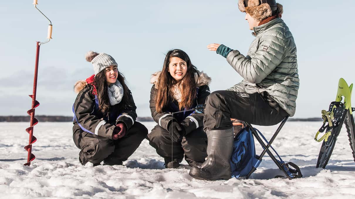 A guide standing beside a red hand ice auger, talking to two guests on fishing stools