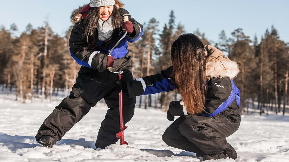 Two women in winter overalls laughing as they hold short ice fishing rods on a frozen Lapland lake