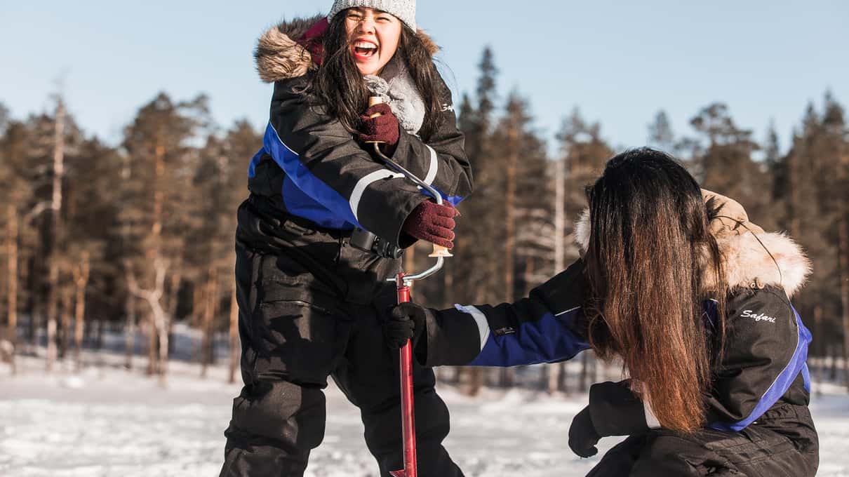 Two guests in fur-trimmed winter overalls laughing as they jig their lines on a sunny frozen lake