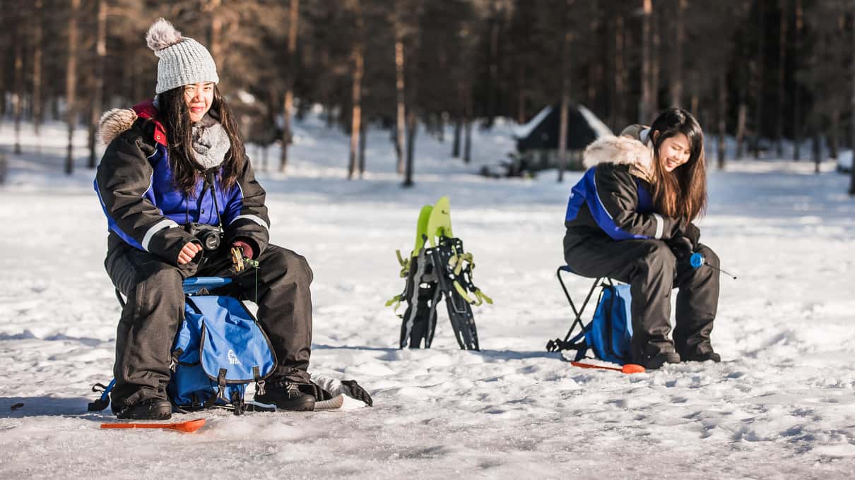 Guests on folding stools fishing through holes on a frozen lake in front of a pine forest