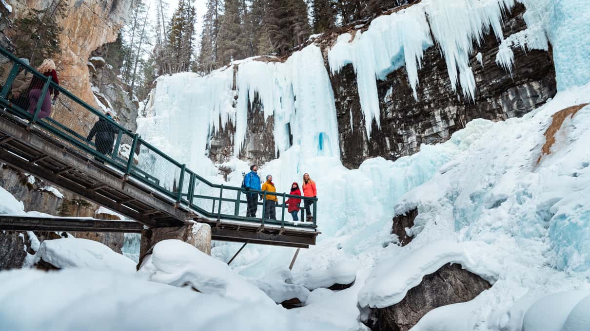 A small group on a steel catwalk below a tall wall of blue frozen waterfall in Johnston Canyon