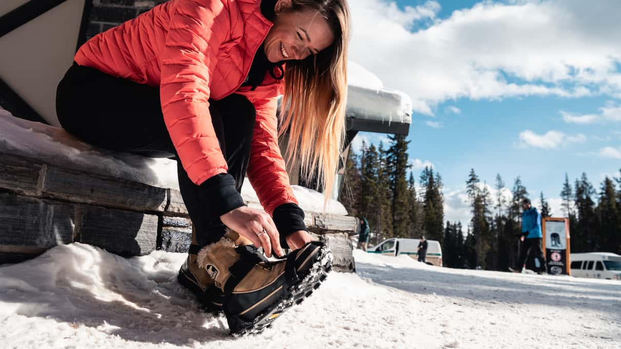 Hiker sitting on a wooden bench pulling ice cleats over her winter boots under pines and blue sky