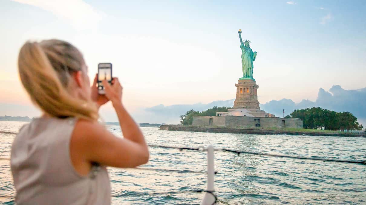 Woman taking a phone photo of the Statue of Liberty from the deck of a ferry in soft evening light