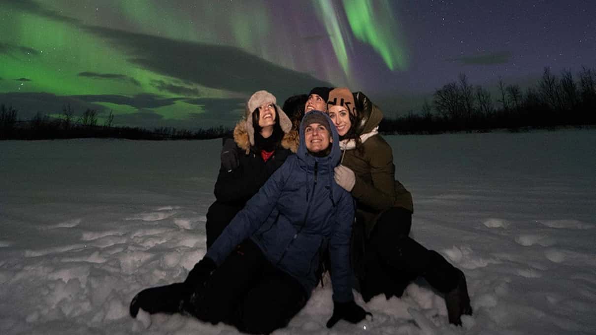 A couple standing close together in the snow for a photo with a green aurora glowing behind them