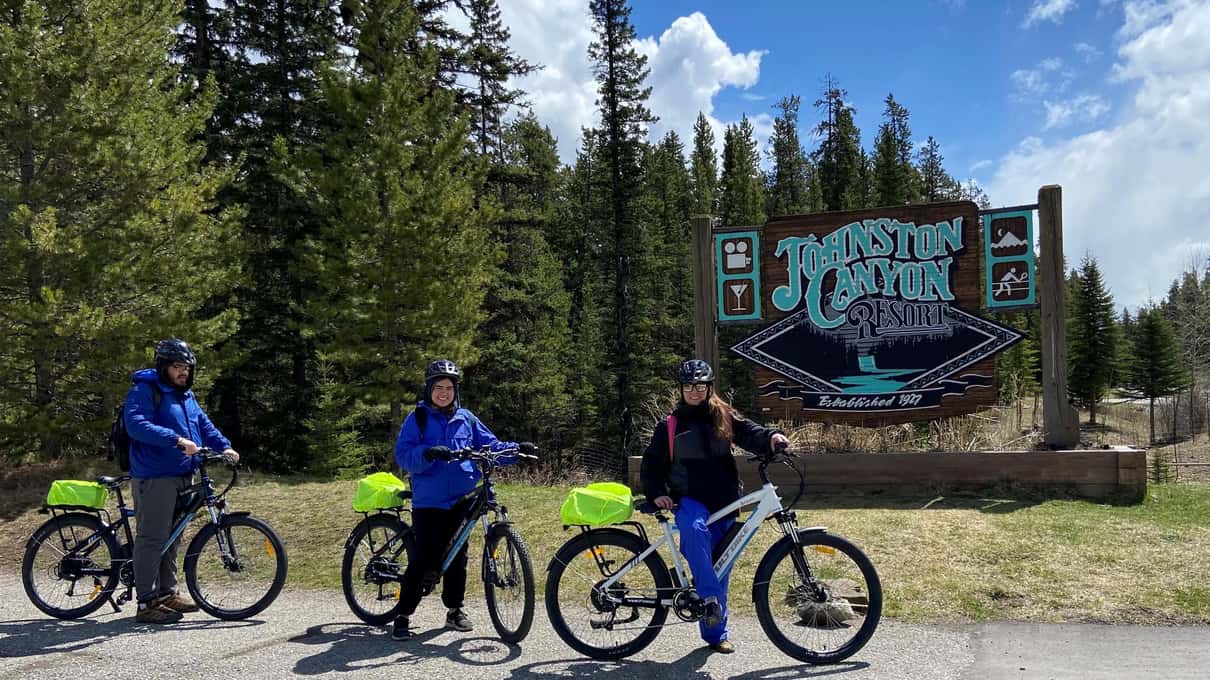 Three riders standing with their e-bikes beside the wooden Johnston Canyon sign in summer