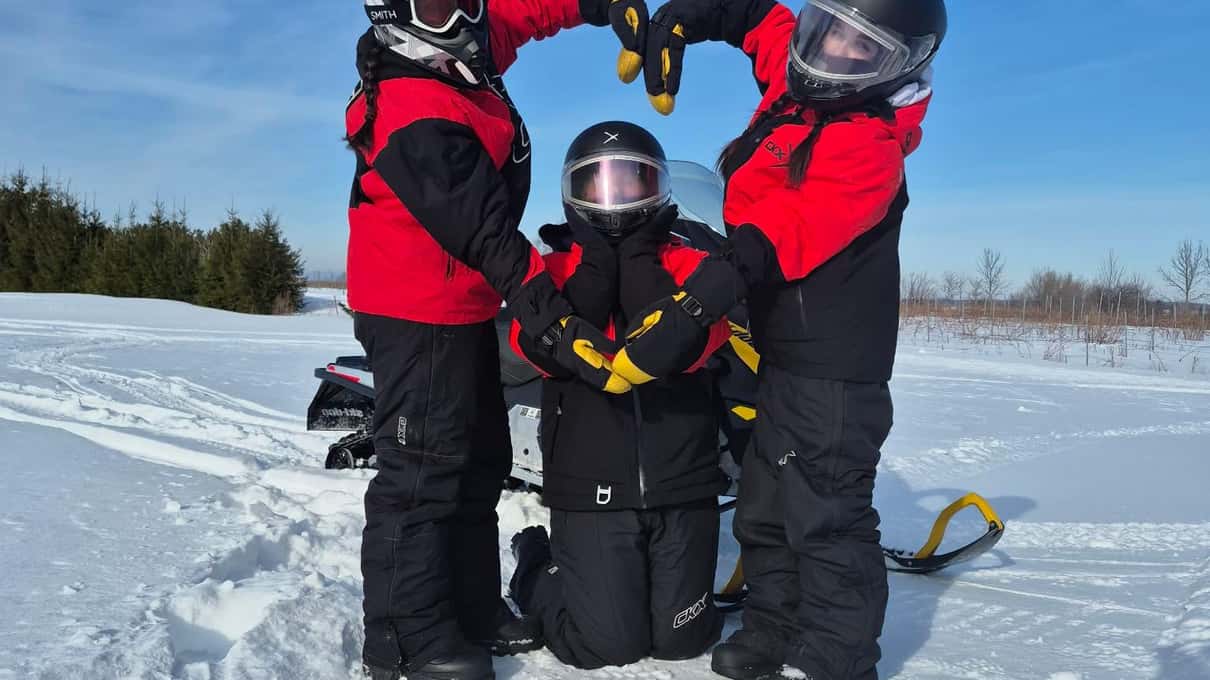 Three snowmobilers in red suits and helmets making a heart shape with their arms in the snow