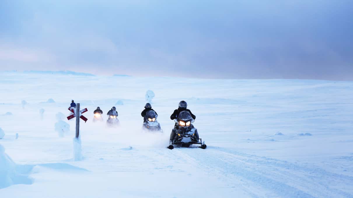 A line of snowmobiles with headlights on, crossing open snow past a trail marker