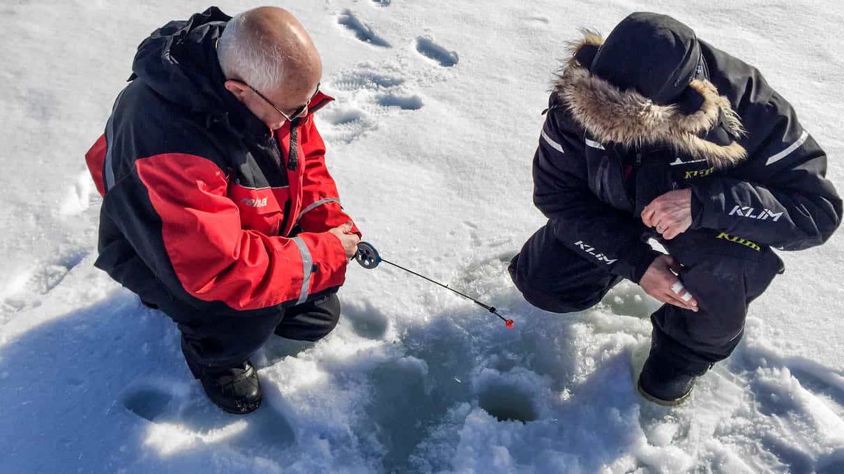 A guide in a red jacket showing a visitor how to fish through a hole in the ice