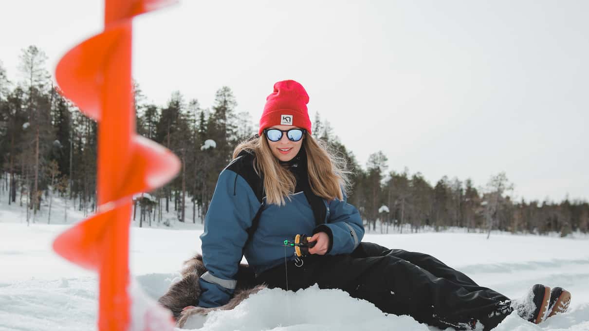 A woman in a red beanie sitting on the snow beside an orange ice auger on a frozen lake