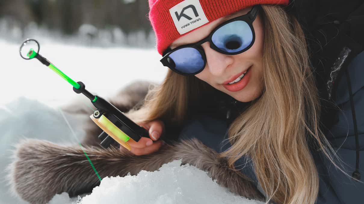 A smiling angler in sunglasses holding a small green ice fishing rod on a frozen lake