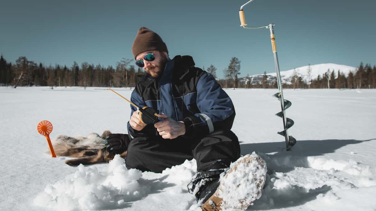 A man fishing on a reindeer skin beside an ice auger with the Levi fell on the horizon