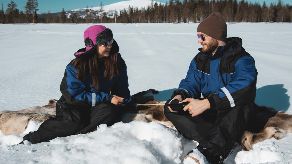 Two people in winter overalls sitting on reindeer skins on the ice while fishing through a hole