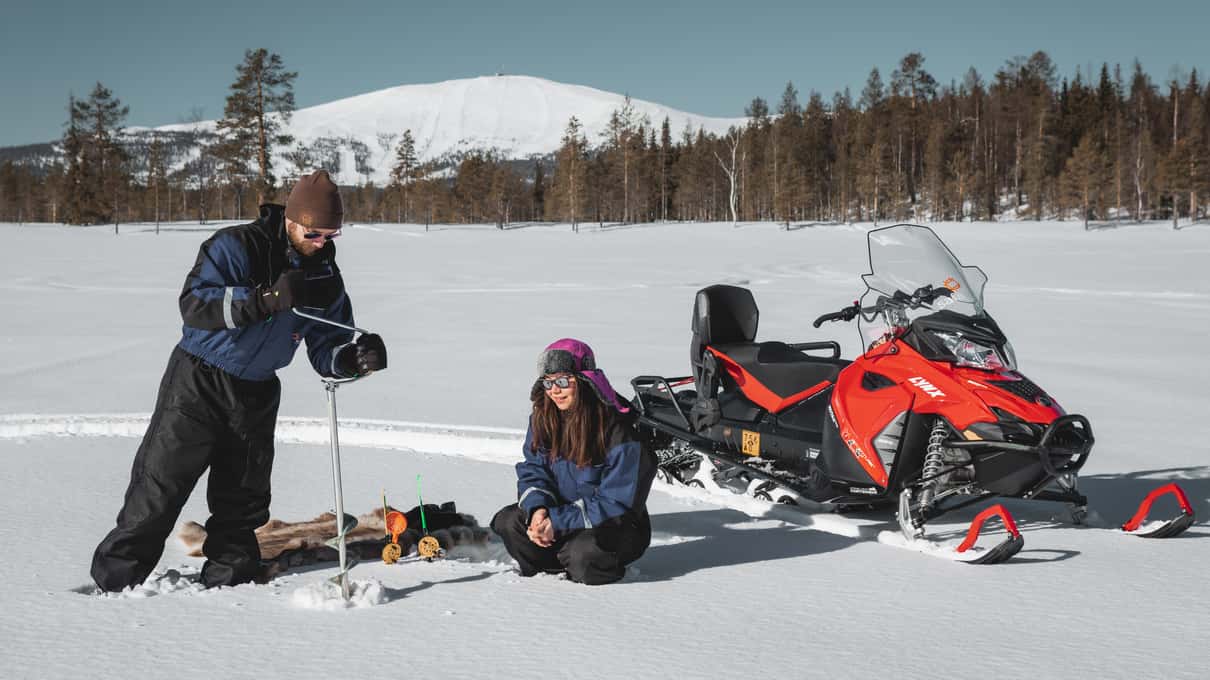 A guide drilling a hole in lake ice beside a red snowmobile, with a snowy fell behind
