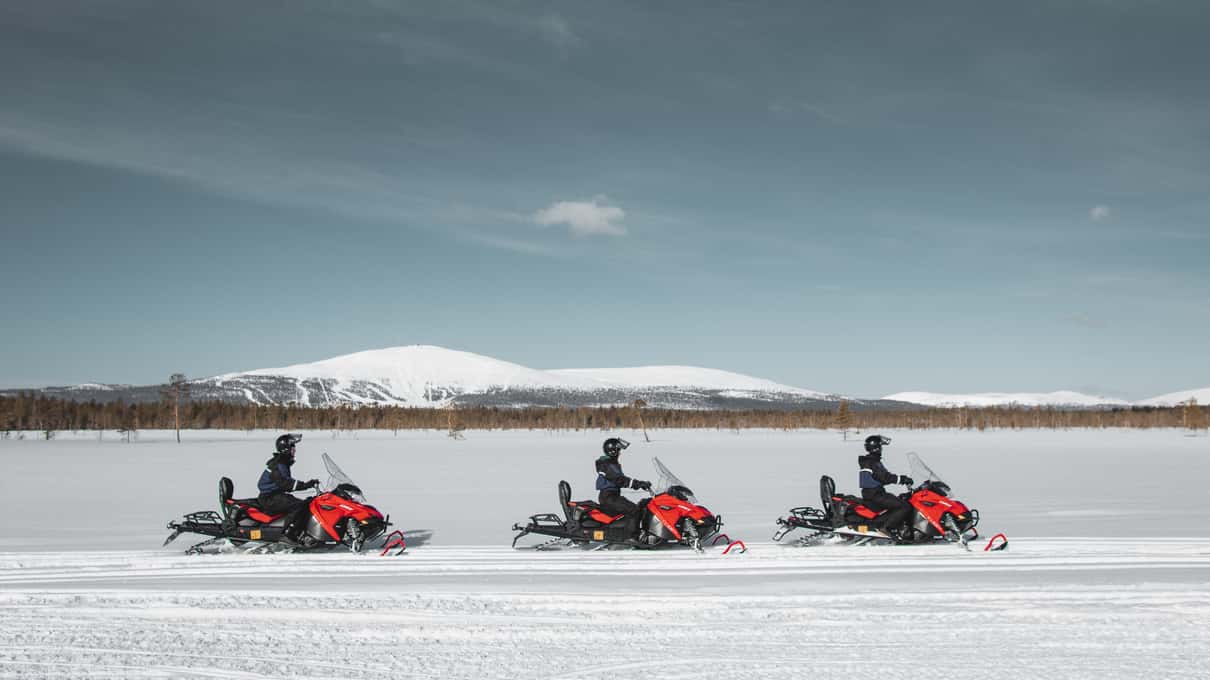 Three red snowmobiles crossing a frozen lake with the rounded Levi fell behind them