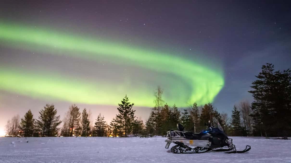 A green aurora arc over parked snowmobiles in the dark on an Arctic Circle Snowmobile Park evening ride