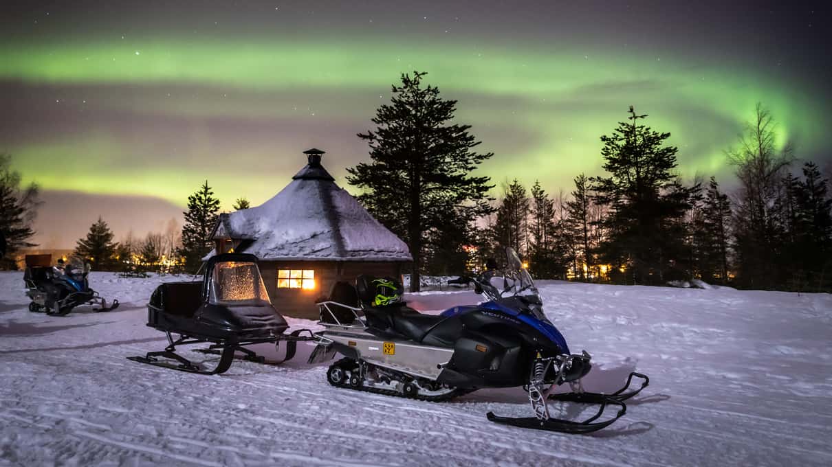 Green northern lights over a wooden hut in the forest, with snowmobiles parked in front