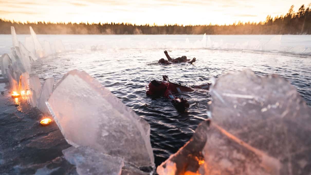 Two floaters in rescue suits lying in dark lake water between lit blocks of ice at night