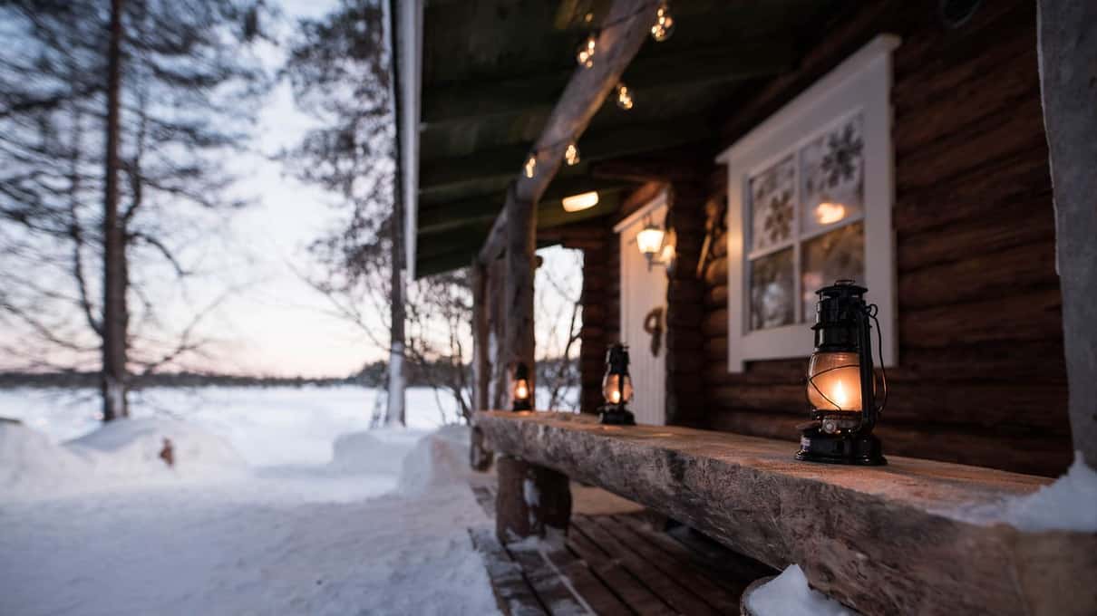 A snow-covered log cabin porch lit by lanterns beside a frozen lake